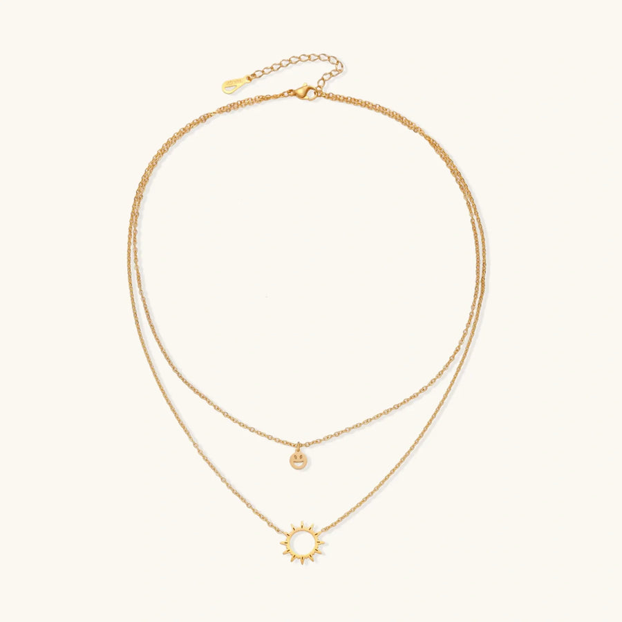 Gold layered necklace set with sunburst and smiley face pendants on delicate chains against white background.