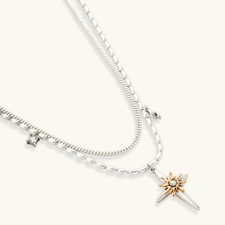 Layered silver-toned necklace set with cross pendant and gold sun charm detail, matching drop earrings on white background.