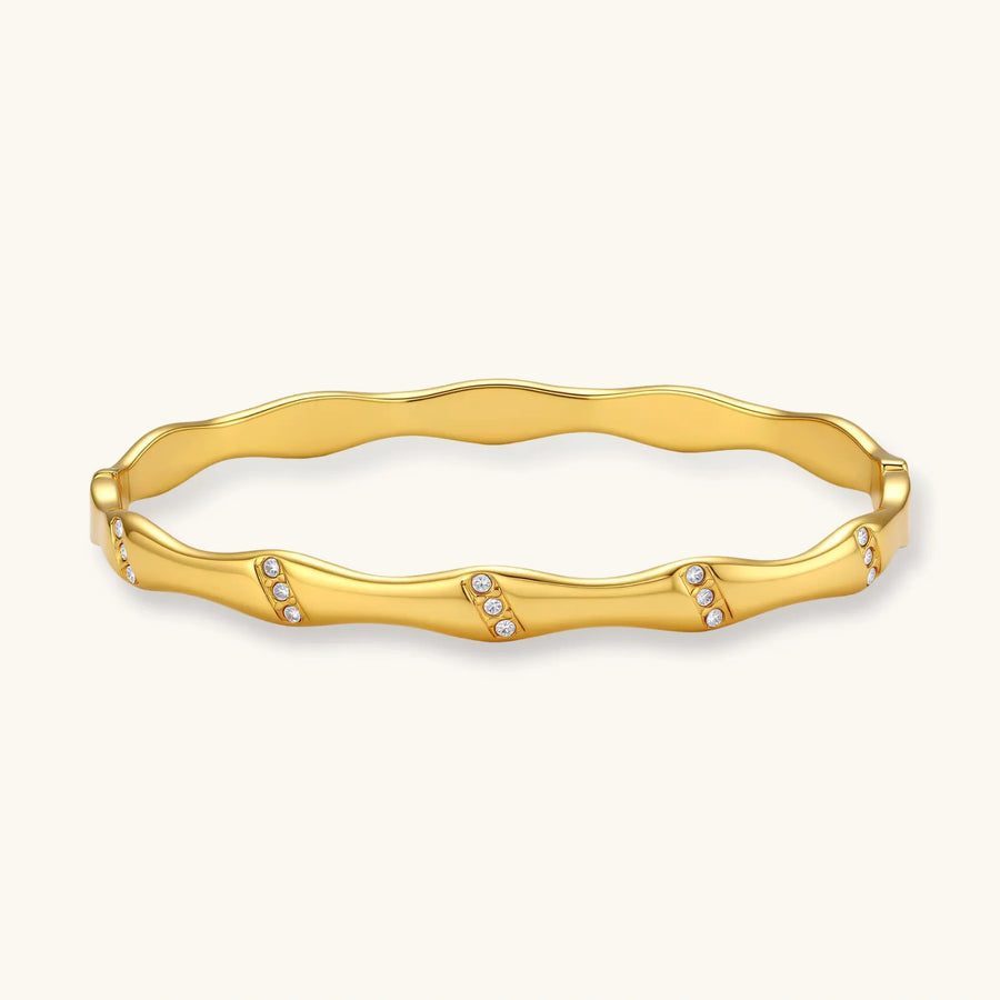Gold-toned wavy bangle with bamboo-style segments and CZ crystal clusters on cream background. 18K plated stainless steel.