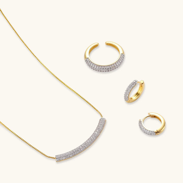 Gold-plated curved necklace, hoop earrings, and adjustable ring with pavé cubic zirconia stones on cream background.