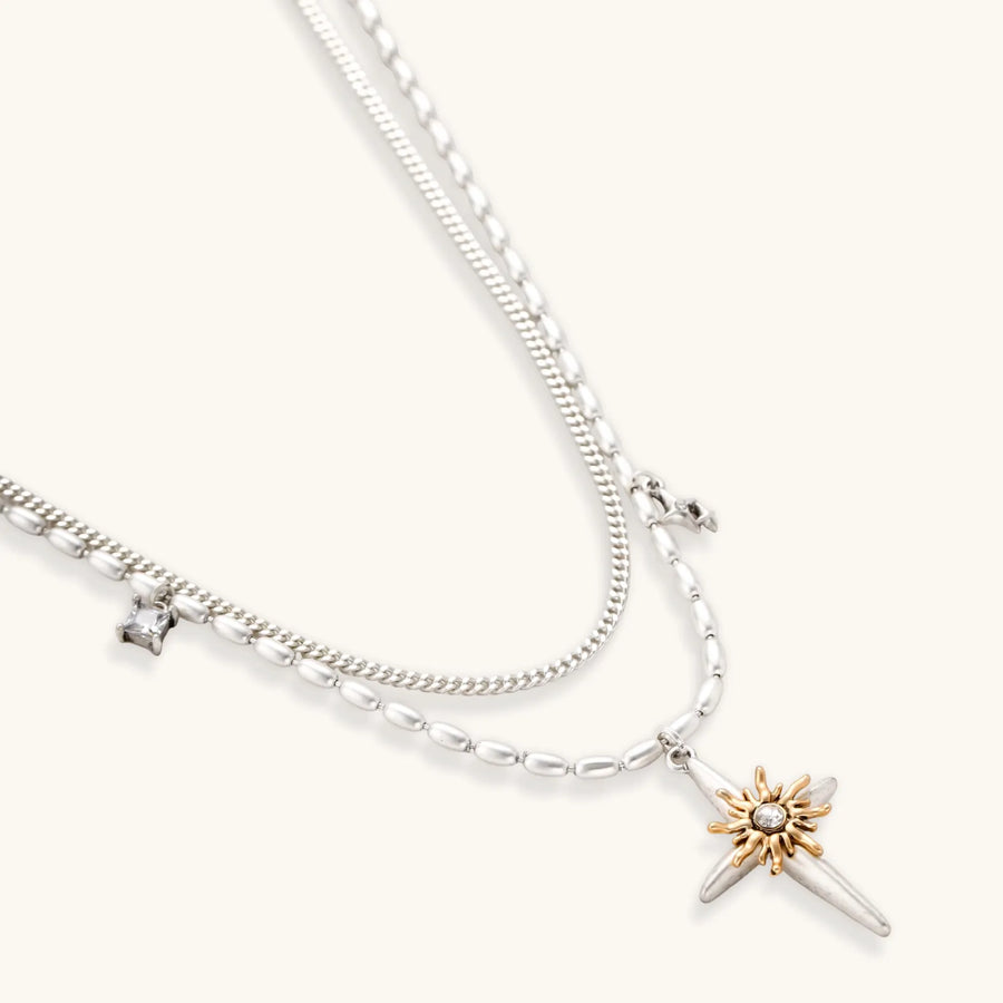Two-layer silver-toned necklace with star charms and dagger pendant featuring golden sun motif on white background.