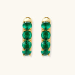 Close-up of gold-plated huggie hoop earrings featuring four emerald green cubic zirconia stones on white background.