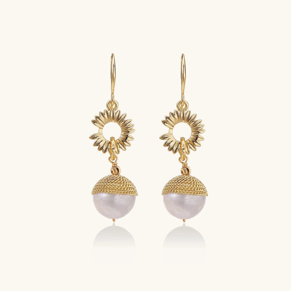 Gold sunburst charm earrings with baroque pearl drops on French hooks against cream background