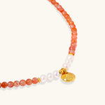Beaded bracelet with coral-orange carnelian stones, white pearls, and gold heart charm on cream background