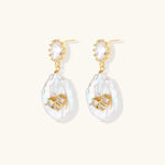 Gold-plated drop earrings with iridescent baroque pearls and crystal flower accents on cream background