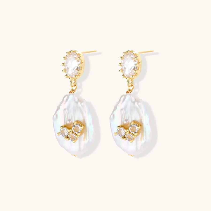 Gold-plated drop earrings with iridescent baroque pearls and crystal flower accents on cream background