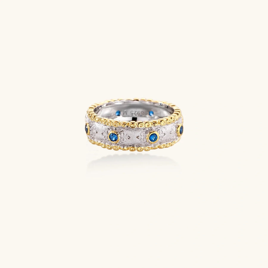 Two-tone ring with brushed silver center, gold beaded edges, and blue zirconia stones on white background.
