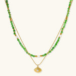 Layered necklace with green stone beads, gold spacers, white accents, and protective eye pendant on cream background.