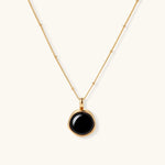 Circular black agate pendant on gold-plated sterling silver chain against cream background, showing modern minimalist design.