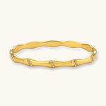 Gold-toned wavy bangle with bamboo-style segments and CZ crystal clusters on cream background. 18K plated stainless steel.